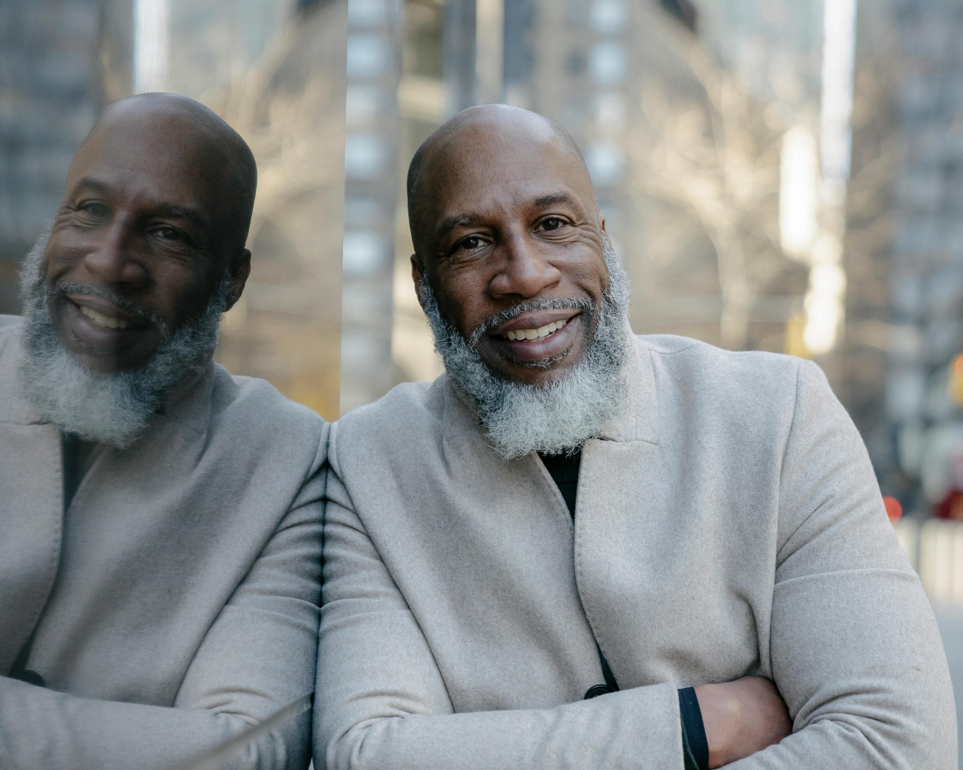 Black man with a gray beard smiling in a light gray coat, outdoors, leaning on a building that shows his mirrored reflection.